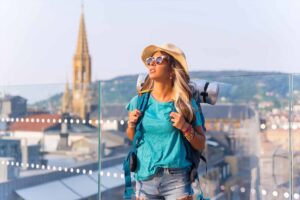 Junge Frau mit Sonnenbrille und Rucksack blickt lächelnd über eine europäische Altstadt.